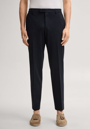 Man draagt zwarte, op maat gemaakte broek en beige suède loafers met kwastjes, staand tegen een effen lichte achtergrond.