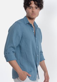 Camicia in lino azzurro chiaro con colletto a bottoni, maniche rimboccate e vestibilità comoda. Presenta bottoni bianchi e una trama sottile.