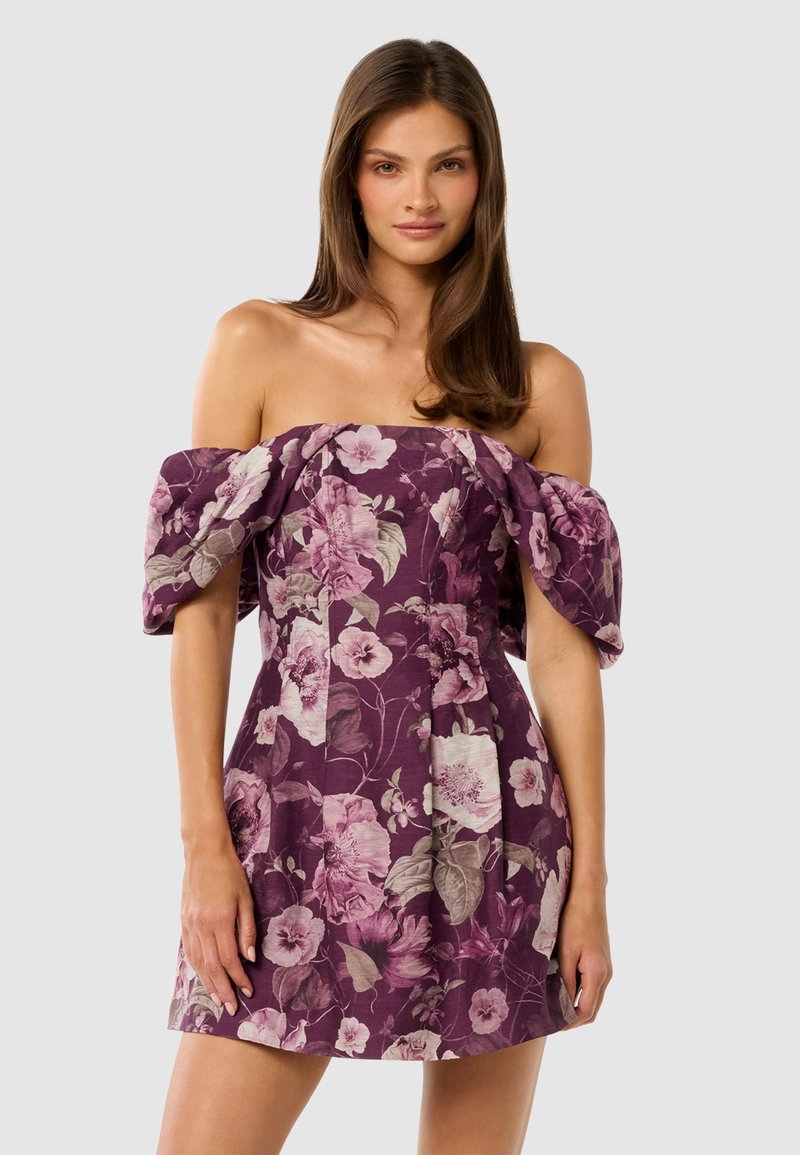 Vrouw met lang bruin haar in een korte, off-shoulder paarse jurk met roze en beige bloemenpatronen, staande tegen een effen achtergrond.