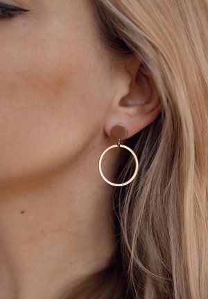 Gold-tone circular hoop earrings with a smooth finish, featuring a smaller round post attachment, showcased beside long, straight hair.