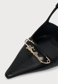 Chaussure noire à bout pointu avec un détail épingle à nourrice dorée affichant un nom de marque en écriture cursive sur la bride avant.