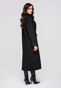 Svart lång kappa av texturerat tyg med hög krage och sidospjäll, kombinerad med knähöga svarta klackstövlar.