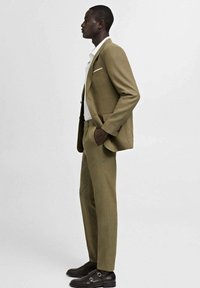 Khaki suit with a tailored fit, featuring a single button closure, a white shirt, and dark brown shoes with a buckle accent.