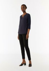 Marineblaues Oberteil mit dreiviertel Ärmeln und V-Ausschnitt, kombiniert mit schwarzen Slim-Fit-Hosen und schwarzen High-Heel-Mules, vor einem weißen Hintergrund.