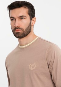 Langärmliges beiges Hemd aus weichem Stoff, mit einem runden Halsausschnitt mit cremefarbener Einfassung und einem kleinen gestickten Logo auf der Brust.