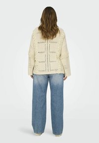 Krem pulover iz pletene tkanine z odprtim vzorcem, frufru robovi in oversized rokavi, v kombinaciji z širokimi modrimi kavbojkami.