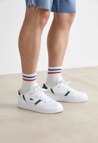 White leather sneakers with green accents, textured sides, and perforations. Paired with white socks featuring red and blue stripes.