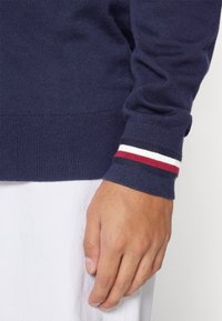 Maglione blu navy con polsini a coste. I polsini presentano strisce rosse, bianche e blu navy. Texture morbida e design aderente. Mano visibile appoggiata sul tessuto.