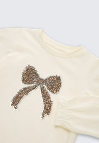 Crème-kleurig shirt met lange mouwen, voorzien van een grote pailletten strikontwerp in metallic tinten van brons en zilver aan de voorkant.