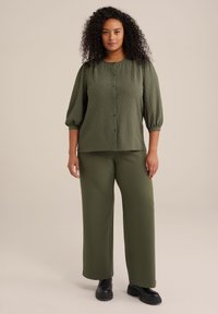 Blusa verde oliva con pois testurizzati, chiusura a bottoni e maniche a tre quarti abbinata a pantaloni larghi coordinati e stivali neri spessi.