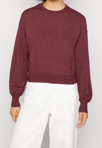 Osoba nosząca bordowy sweter z długim rękawem i okrągłym dekoltem, zestawiony z białymi spodniami o wysokim stanie, stoi na jednolitym tle.