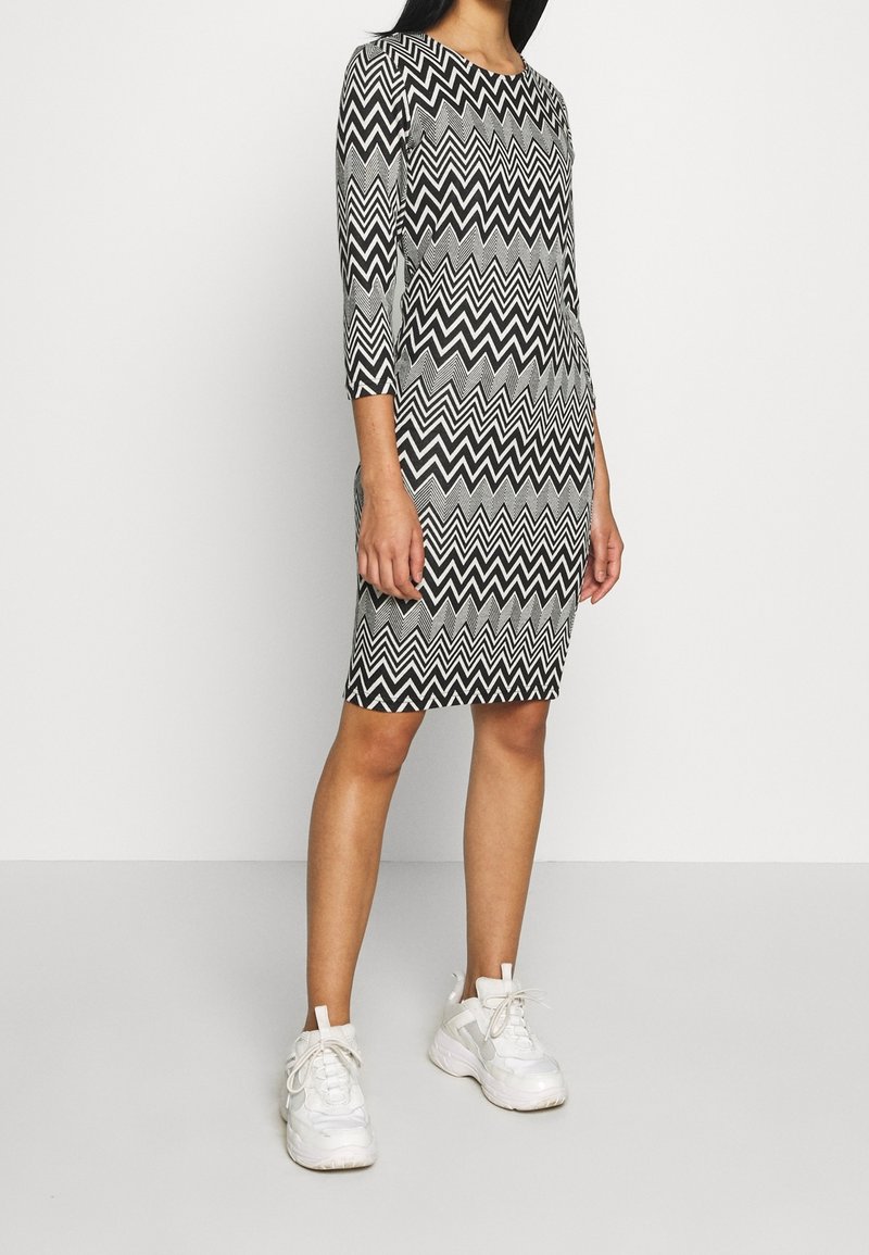 Femme portant une robe noire et blanche à motif zigzag, longueur genoux, manches trois-quarts et baskets blanches épaisses.