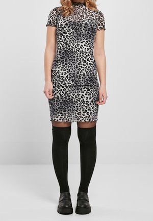 Žena v krátkých rukávech, leopardím vzorem na šatech, černé podkolenky a černé platformové boty, stojící před jednoduchým pozadím.