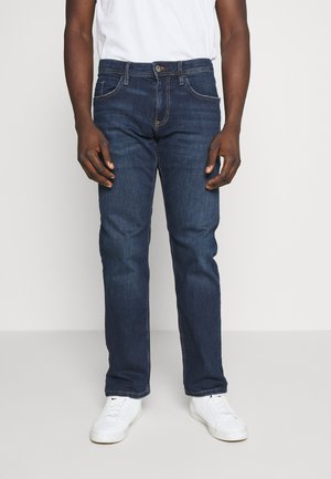 Homme portant un jean droit bleu foncé, des baskets blanches et un t-shirt blanc, debout devant un fond clair uni.