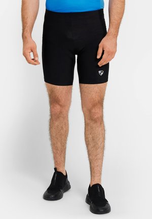 Man wearing black fitted shorts, black slip-on sneakers, and a blue top, standing against a plain light background.