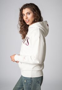 Witte hoodie met een voorzak, trekkoord capuchon en wijnrode letters. Kenmerkt ribgebreide mouwen en onderkant, gecombineerd met vervaagde blauwe jeans.