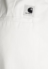 Tissu blanc avec une poche rectangulaire cousue et un petit patch logo Carhartt comportant un symbole de vague noir et le nom de la marque.