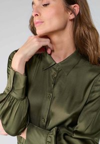 Blouse en satin vert olive avec un col classique, un devant boutonné et des manches bouffantes avec des accents de boutons. Texture lisse et coupe ajustée.