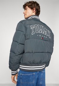 Chaqueta de pluma gris con letras blancas "TOMMY JEANS" en la parte posterior, con puños y dobladillo acanalados con rayas blancas y grises.