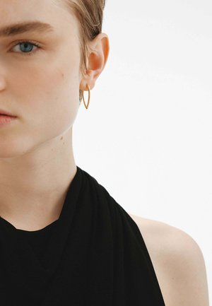 Pendiente de aro dorado exhibido sobre una blusa de tela negra, con un primer plano del rostro de un modelo con piel clara y cabello corto y desaliñado.