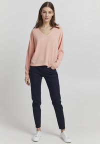 Helles rosa, strukturiertes V-Ausschnitt-Pullover mit fallenden Schultern, kombiniert mit navy blauen Slim-Fit-Hosen und weißen Sneakers.
