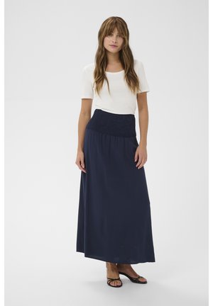 Kvinde med langt brunt hår iført en hvid kortærmet top, navyblå maxinederdel med høj talje og sorte åbne hælede sandaler.