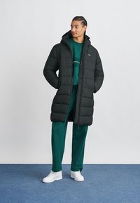 Μαύρο πούφο μπουφάν με κουκούλα, έχει οριζόντια ραφή, μπροστινό φερμουάρ και δύο πλευρικές τσέπες. Φορεμένο πάνω από πράσινο σύνολο και λευκά αθλητικά παπούτσια.