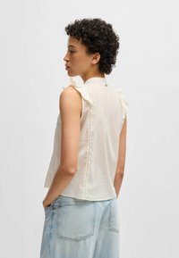 Ärmlös, off-white blus med volangdetaljer vid axlarna, har hög krage och en knappdetalj på baksidan, med subtila sömmar.