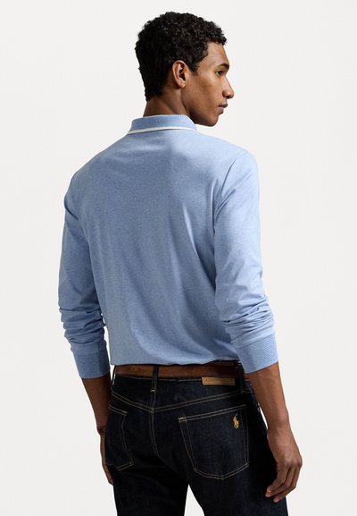Gai zils garām piedurknēm polo krekls ar apkakli. Modelis valkā tumšas džinses ar brūnu ādas jostu. Iezīmē minimālas šuvju detaļas un logo.