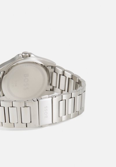 Montre en métal argenté avec un bracelet à maillons, un boîtier rond et une finition polie. L'arrière présente une surface lisse avec le nom de la marque gravé.