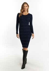 Marineblaues Rippstrickkleid mit langen Ärmeln, das mit dekorativen weißen Perlen versehen ist, ergänzt durch eine körpernahe Silhouette und kniehohe schwarze Stiefel.