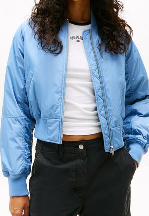 Femme portant une veste bomber bleu clair courte sur un t-shirt blanc Tommy Hilfiger et un pantalon noir taille haute, les mains dans les poches.