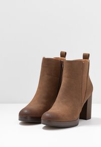 Bottines en fausse suède marron avec un bout arrondi, fermeture éclair latérale et talon empilé. Présente des accents de couture et des languettes de tirage sur le col.