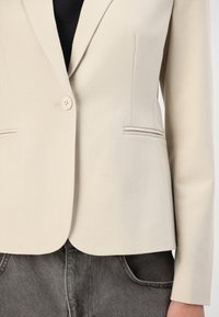 Beige bleiseri valmistettu sileästä kankaasta, jossa on yksi napitus, hapsulaatikot ja kaksi etutaskua. Yhdistetty harmaisiin farkkuihin.