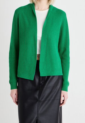 Femme portant un cardigan en maille côtelée verte sur un crop top blanc et une jupe en cuir noire sur un fond blanc uni.