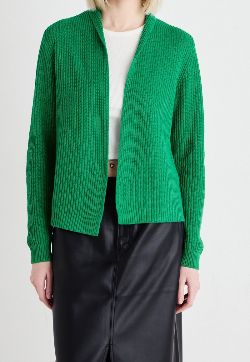Femme portant un cardigan en maille côtelée verte sur un crop top blanc et une jupe en cuir noire sur un fond blanc uni.