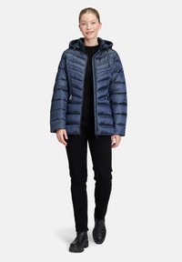 Marineblauwe pufferjack met capuchon, voorzien van een gequiltde textuur, ritssluiting aan de voorkant en zijzakken. Draag het over een zwarte trui en broek.
