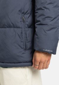Mornarica puffer jakna s kvadratnim dizajnom, bočnim džepom sa zipom i Quiksilver logom. Glatka tkanina s mekim teksturom, uski manšeti.
