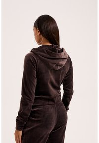 Femme aux longs cheveux foncés portant un sweat à capuche en velours marron foncé et un pantalon assorti avec des logos cloutés, de dos sur fond neutre.