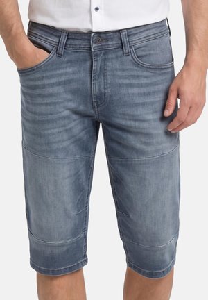 Mann trägt ausgewaschene blaue Denim-Shorts mit Fronttaschen, sichtbaren Nähten und ein weißes Hemd, das eingesteckt ist, Hände entspannt seitlich.