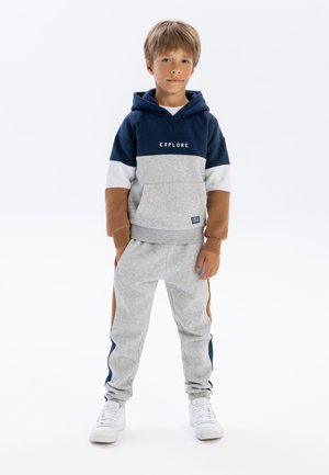 Marineblauer und grauer Kapuzenpullover mit dem Schriftzug "EXPLORE", ergänzt durch graue Sweatpants mit braunen Akzenten und seitlichen Streifen. Weiße Sneaker.