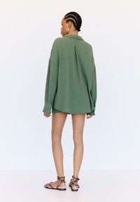 Grüne, übergroße Hemdbluse aus strukturiertem Stoff, mit langen Ärmeln und einem Faltenaufsatz hinten. Kombiniert mit braunen Riemchensandalen.