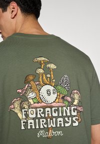 Μπλούζα από βαμβάκι σε ελιά πράσινο χρώμα με πολύχρωμο γραφικό μανιταριού και το κείμενο "FORAGING FAIRWAYS" με έντονα γράμματα στην πλάτη.
