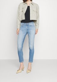 Avkortad, beige rutiga jacka över en svart linne, i kombination med ljusblå skinny jeans och beige klackskor med dekorativa detaljer.