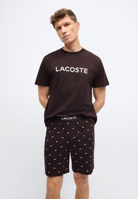 Νεαρός άνδρας που φοράει μαύρο t-shirt Lacoste με λευκό λογότυπο και αντίστοιχα μαύρα σορτς με μικρά λευκά κροκοδειλάκια, στέκεται σε απλό φόντο.