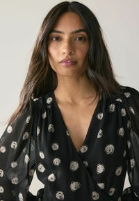 Rochie neagră cu mâneci lungi, transparente și fluide. Prezintă un material texturat și modele circulare albe răspândite pe suprafață.