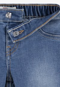 Denim kavbojke s svetlo modrim pranjem, opremljene z kovinskim gumbom, elastičnim pasom in kontrastnimi rumenimi šivi.