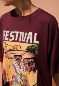 Camiseta de algodón color burdeos con un vibrante gráfico de un hombre con sombrero, con la palabra "FESTIVAL" impresa en letras blancas negritas encima.