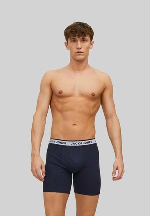 Junger Mann mit kurzen lockigen Haaren, der dunkelblaue Jack & Jones Boxershorts trägt und vor einem grauen Hintergrund steht.