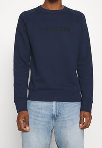 Námořnická modrá mikina s raglánovými rukávy, měkká textura, kulatý límec a černé reliéfní logo "DOCKERS" na přední straně. Diskrétní ribované manžety a lem.